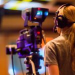 A woman operates a professional camera indoors, focusing on media production.